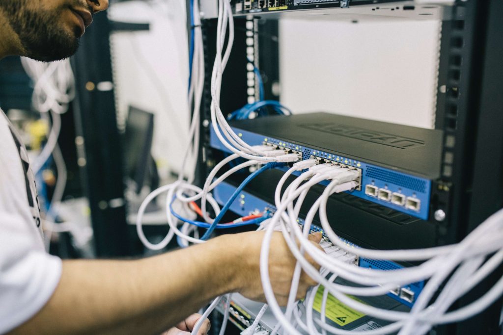 A person works on a server rack, connecting multiple Ethernet cables to network switches and devices. The image shows a close-up of their hands reaching among a cluster of cables, emphasizing the technical nature of the setup.