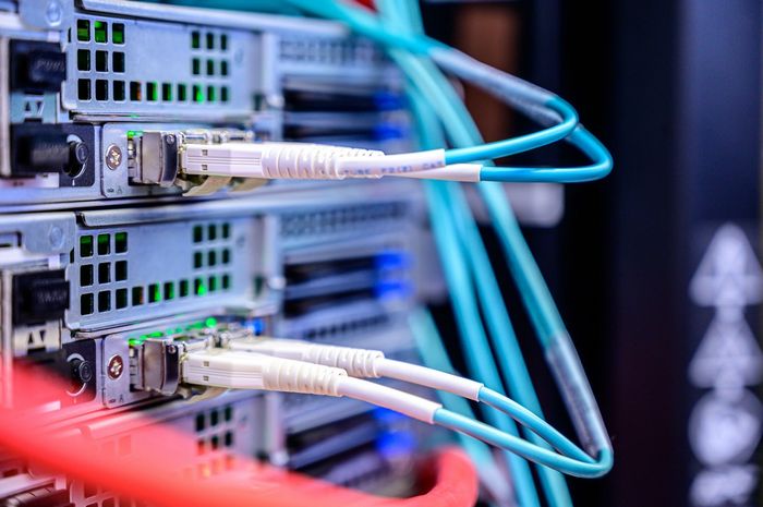 A close-up view of network cables connected to the rear side of server units in a data center. The image features various colored cables, including blue and red, plugged into silver server ports with LED lights indicating activity.