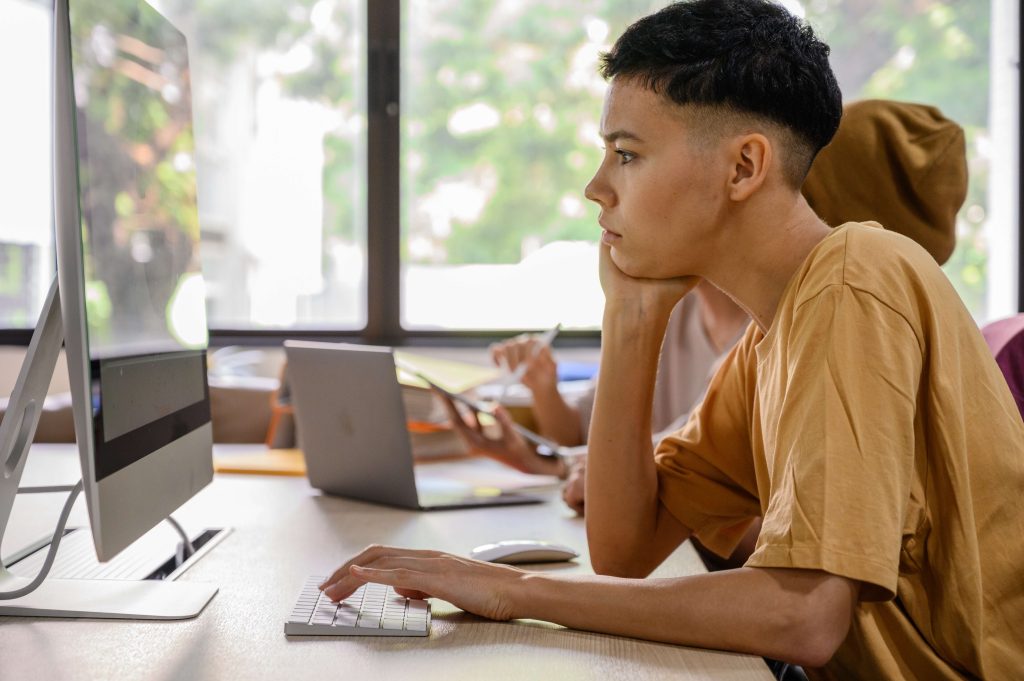 A person with short hair, wearing a brown shirt, is sitting at a desk and focused on a computer screen. They are using a keyboard and mouse. Another person in a hijab is in the background, also working on a laptop. Large windows in the background show greenery outside.