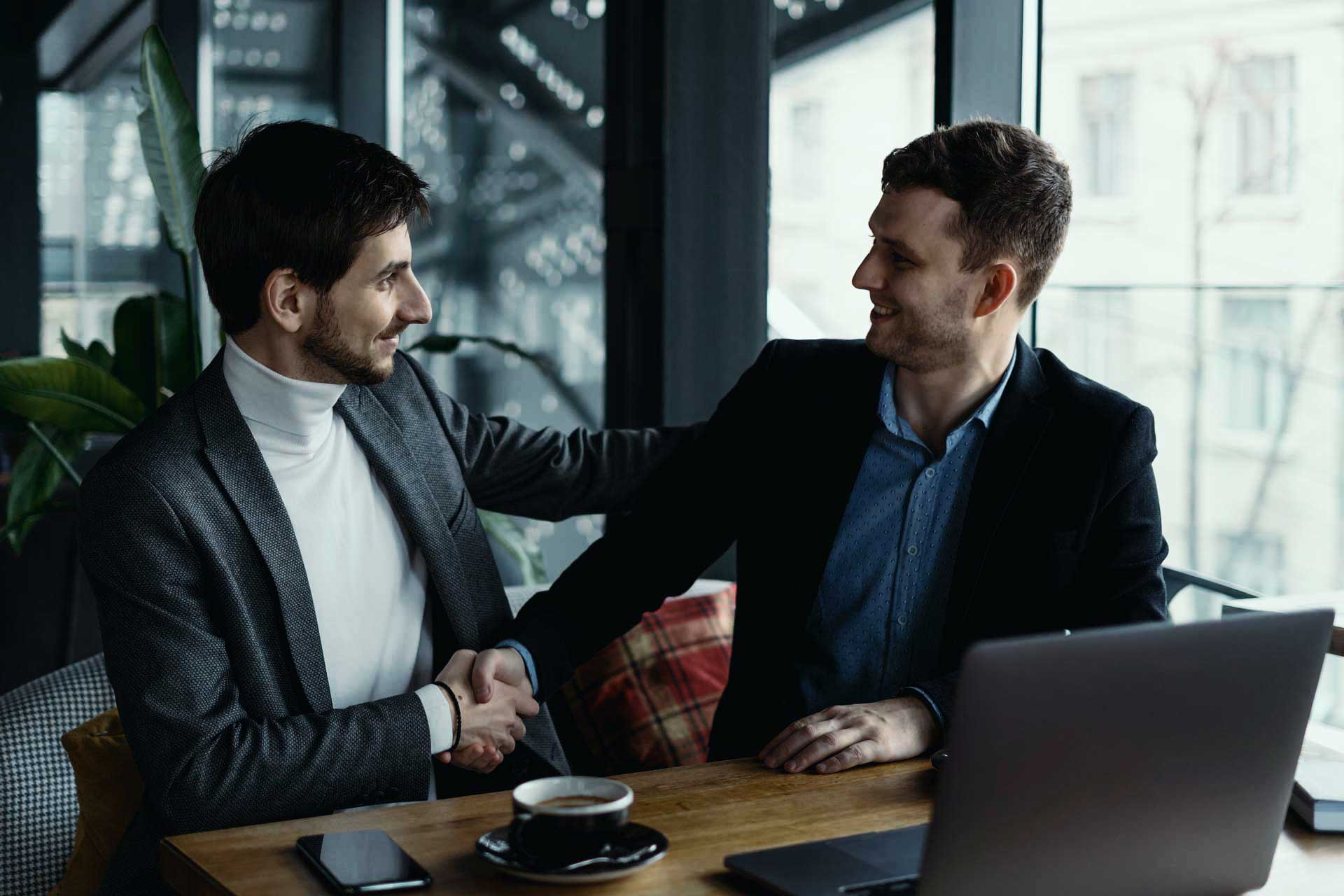 Two men in formal attire are sitting at a wooden table in a modern office, shaking hands and smiling at each other. A laptop, smartphone, and a cup of coffee are on the table. Large windows in the background provide natural light.