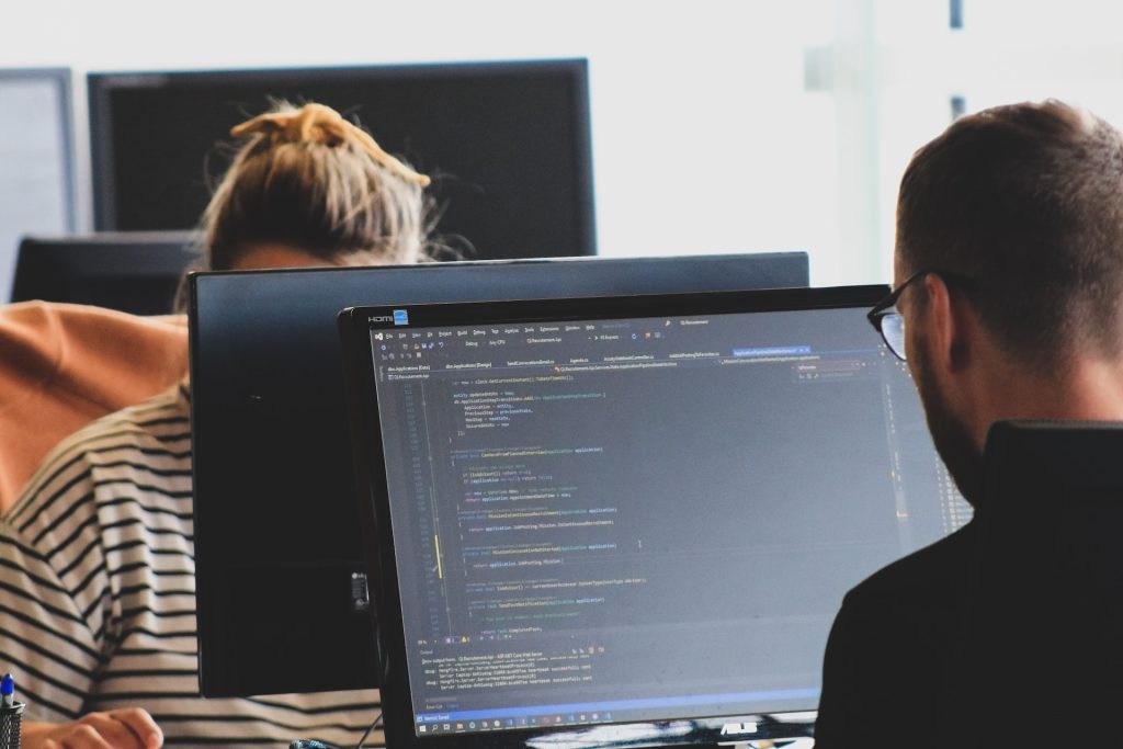 Two individuals are working at computer desks. The person in the foreground is focused on a monitor displaying code in an integrated development environment (IDE), while the person in the background is partially visible, obscured by a screen.