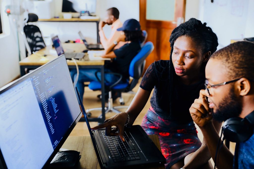 Two people are at a desk, focused on a computer screen displaying code. One person points to the screen while the other looks on, resting a hand on their chin. In the background, another desk with two people working on laptops is visible.