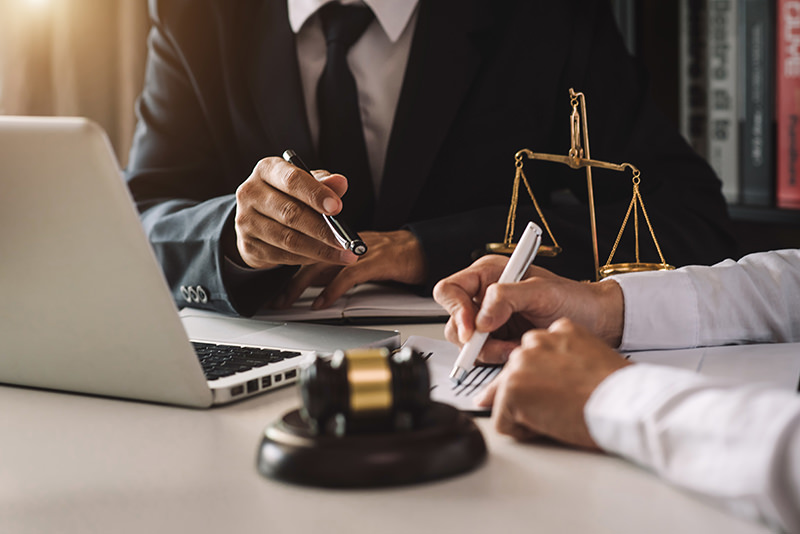 Two people in business attire sit at a desk with a laptop, discussing documents. One person points with a pen, while the other takes notes. A brass scale and a gavel are placed on the desk, suggesting a legal or courtroom setting. Their discussion hints at integrating Managed IT Services into their operations.