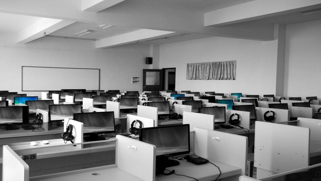 A modern, empty computer lab with multiple rows of workstations. Each desk has a computer monitor, keyboard, and set of headphones. The room has a whiteboard on one wall and large windows with curtains in the back, allowing natural light to enter.