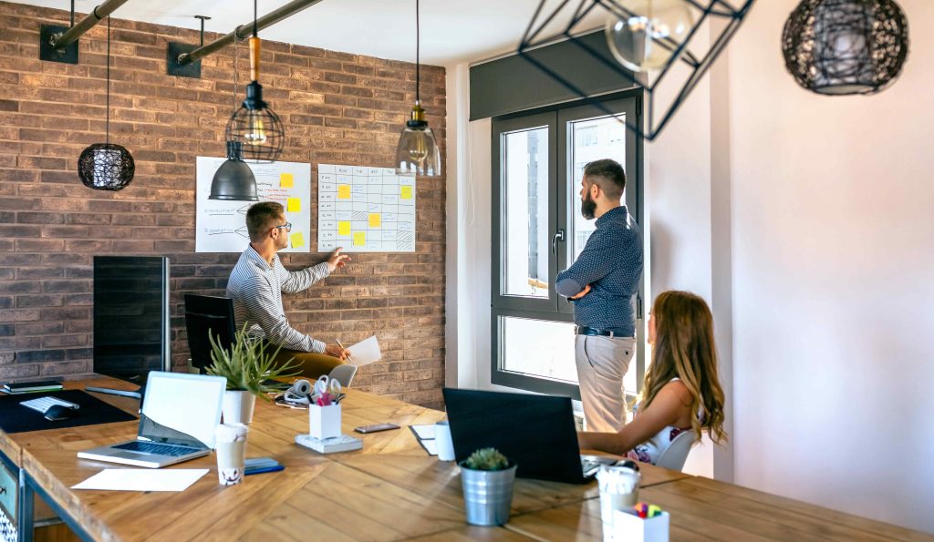 Three people are working in a modern office. One person is seated and pointing to a wall chart while explaining something about managed IT services to two others, one standing by the window and another sitting at the table. The office has brick walls, wooden tables, and hanging light fixtures.