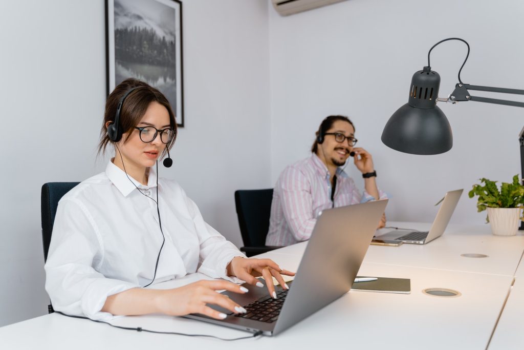 Two people are in an office setting. The person in front is typing on a laptop while wearing a headset, with a focused expression. The person in the background, also wearing a headset, looks at a screen and smiles. A desk lamp and a small plant are on the table.