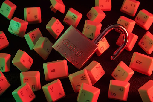 An open padlock is surrounded by scattered keyboard keys against a dark background, illuminated with red and green lighting.