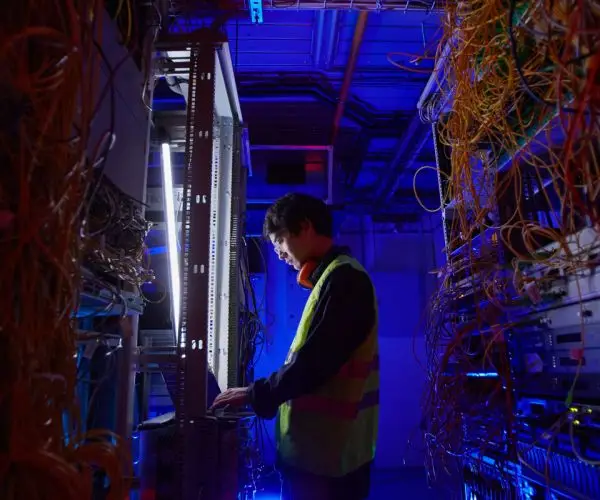 A person wearing a high-visibility vest and headphones is working on a laptop in a dimly lit server room. They are surrounded by racks of servers and tangled cables, illuminated by blue and purple lighting, providing IT consulting services in Seattle.