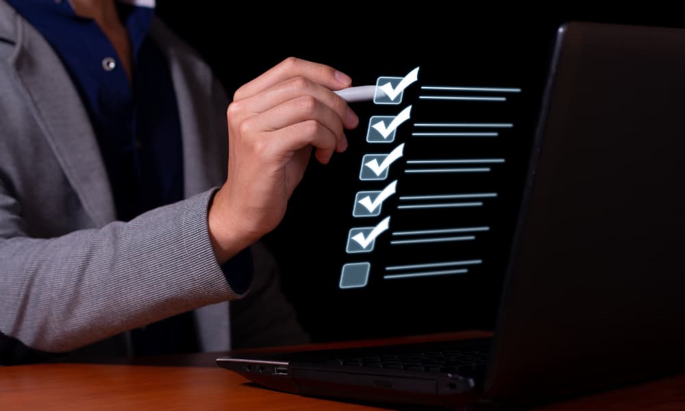 A person in a gray blazer holds a pen, pointing at a holographic cybersecurity assessment checklist projected in front of a laptop screen. The checklist has multiple checked boxes and one empty one. The person is sitting at a wooden surface.