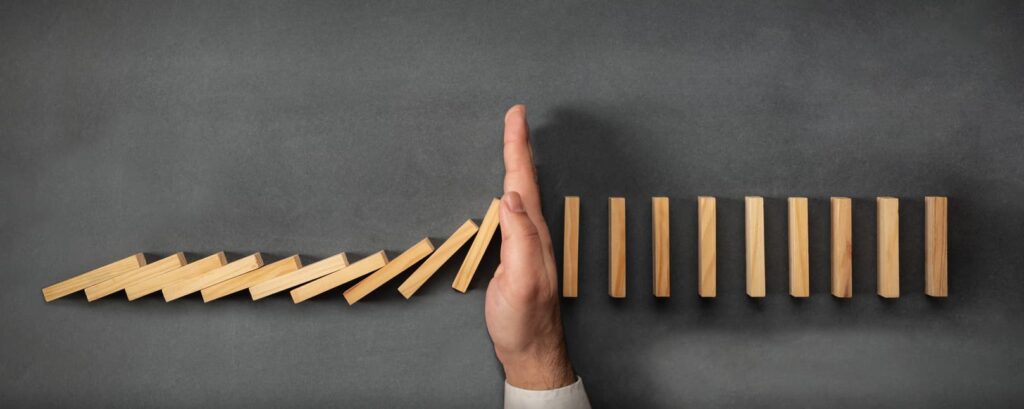 A hand stops falling dominoes, preventing them from toppling others, representing a measure for enhancing security. The backdrop is a chalkboard-like surface, giving a contrast to the wooden blocks and hand.