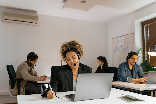 Four people are working in an office, each seated at separate desks with laptops. They are wearing headsets, suggesting an IT Support or Helpdesk scenario. The room has white walls, air conditioning, and various office supplies.