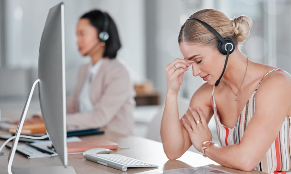 A woman with blonde hair, wearing a striped top and a headset, sits at a desk with a computer, pinching the bridge of her nose in apparent frustration or stress. Coping with VoiP issues, another woman in a blazer and headset works on a computer in the blurred background.